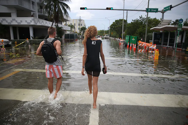A woman in a dress carrying her shoes and a man in red and white striped shorts walk down a street that is filled with water to above their ankles.
