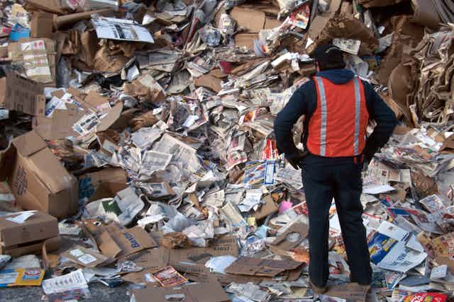 A man in a red hazard vest stands before a cascade of cardboard and newspaper.
