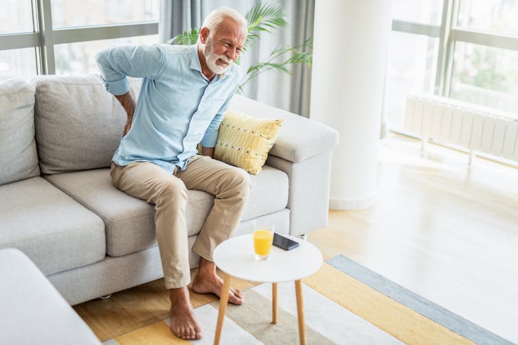 Older man sits on a sofa in his living room and grimaces while holding a hand to his lower back.