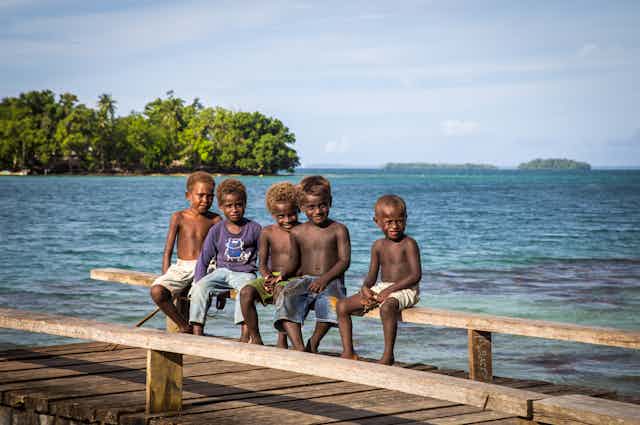 Group of children sitting on a wooden pier in the Solomon Islands, with their backs to the sea.