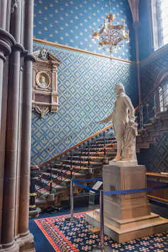 White statue surrounded by a staircase with blue and red carpet, blue and gold wallpapered walls, gold chandelier..