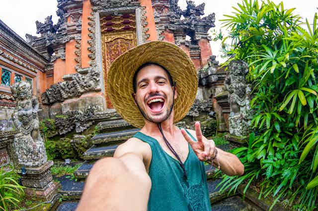 A young man flashes a peace sign and takes a selfie in front of a temple in Bali