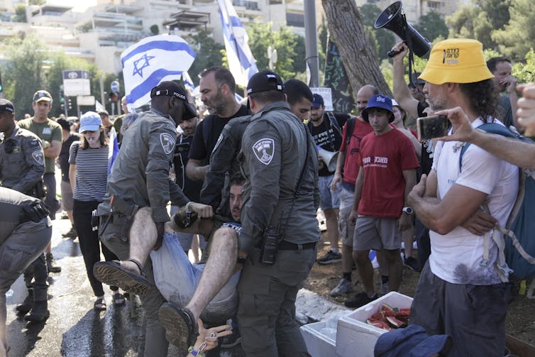 People in uniforms carry an unwilling person away from a group surrounding him.