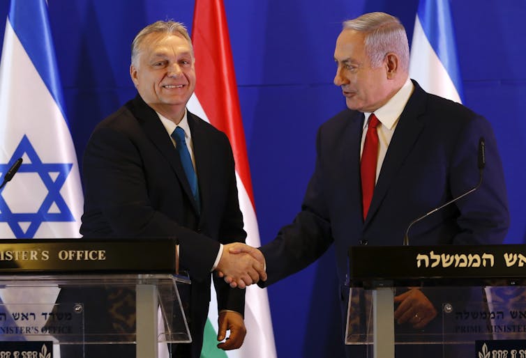 Two men in dark jackets sharking hands in front of flags.