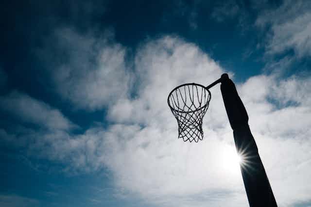 A netball post and hoop set against the backdrop of a cloudy sky, the sun coming through the clouds.