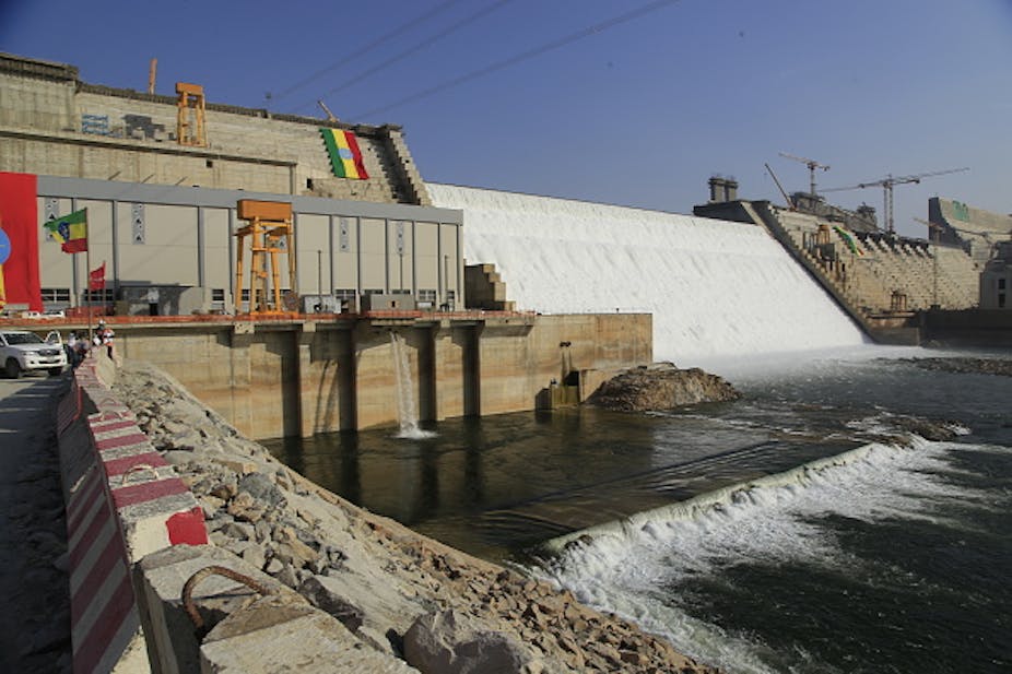A sheet of water falling over a slanted concrete wall into a large pool that is inside concrete walls, with cranes around the walls, and flags with red, yellow and green stripes