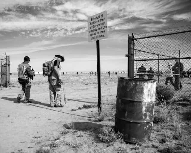 a black and white photograph of two men entering a gated area with a warning sign and a barrel