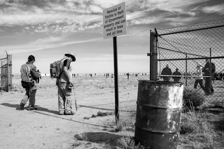 a black and white photograph of two men entering a gated area with a warning sign and a barrel