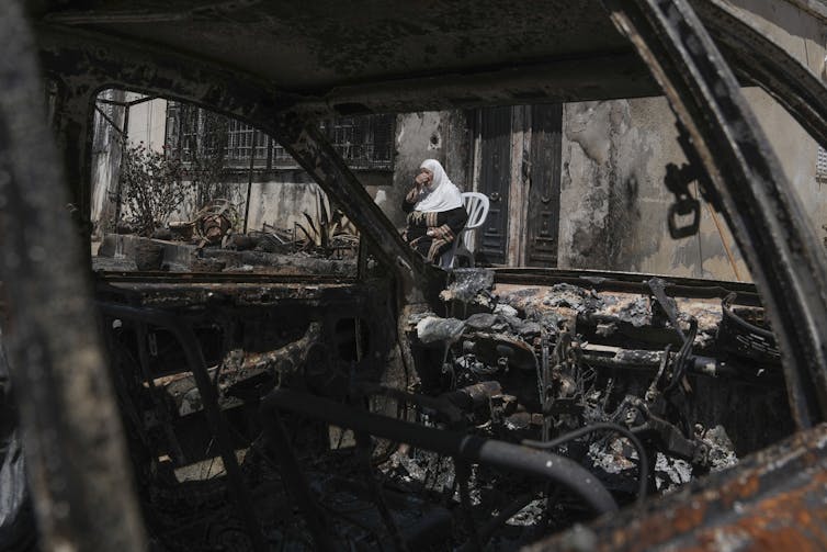 A woman sitting outside a burned-out home, seen through a burned up car.