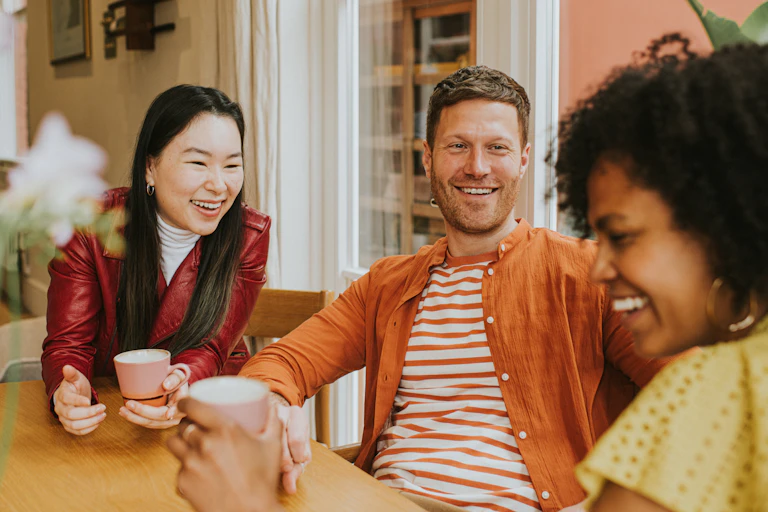 three people with big smiles chat at table with coffee cups