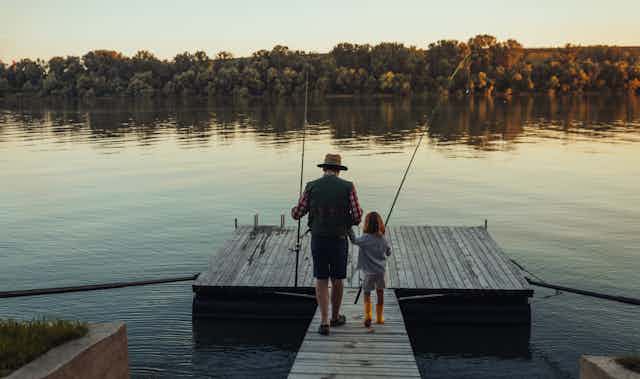 Man and girl hold hands and fishing poles while going fishing together on a dock.
