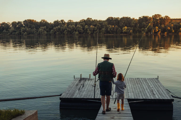 Man and girl hold hands and fishing poles while going fishing together on a dock.