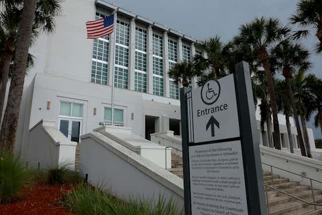 A white building with an American flag is seen from the outside, with palm trees and an entrance sign in front of it.