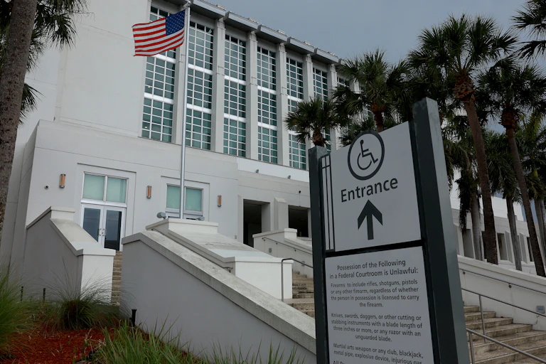 A white building with an American flag is seen from the outside, with palm trees and an entrance sign in front of it.