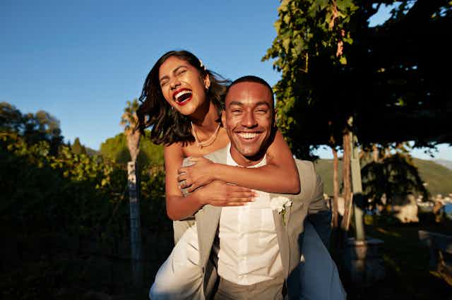 A brunette woman with red lipstick laughs as a man in a suit, smiling, gives her a piggyback ride.