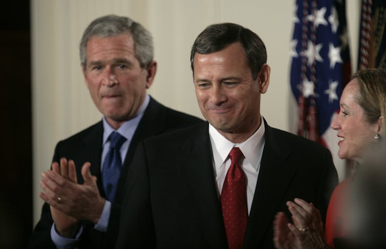 A gray-haired white man is applauding about white man dressed in a business suit as his wife looks on approvingly.