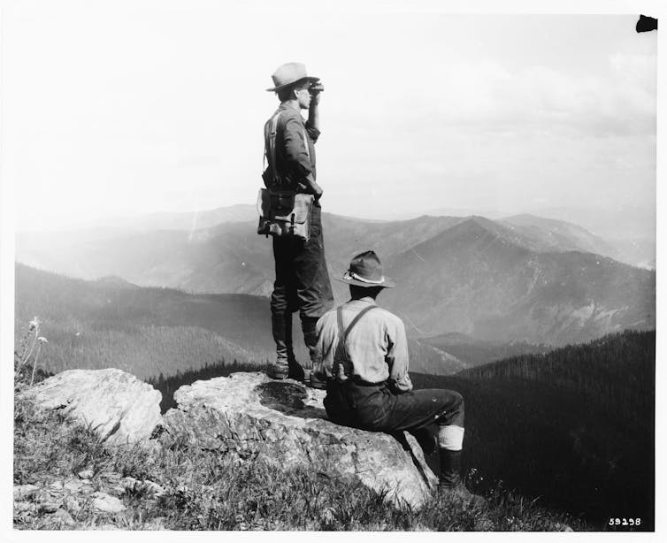 A black and white photo shows a man standing on a mountaintop rock looking through binoculars, with mountains in the background. Another sits on the rock beside him.
