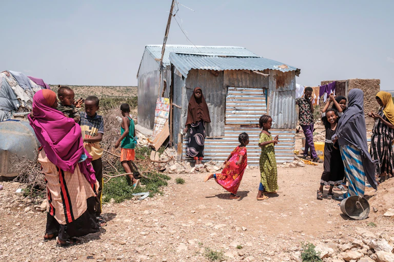 A group of women and children are pictured standing in front of and next to a makeshift home that is a dilapidated zinc structure
