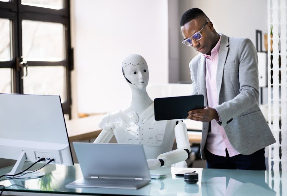 A young man in an office shows an ipad to a human-like, white plastic robot, sitting at a desk and working on a computer