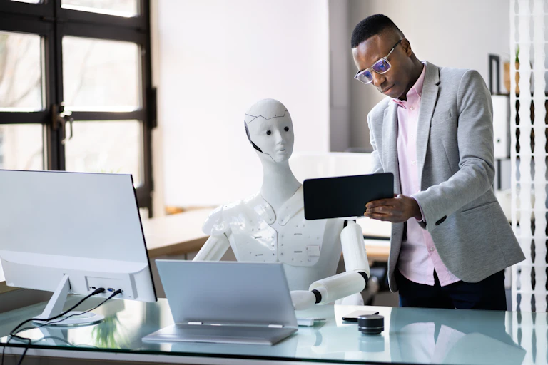 A young man in an office shows an ipad to a human-like, white plastic robot, sitting at a desk and working on a computer