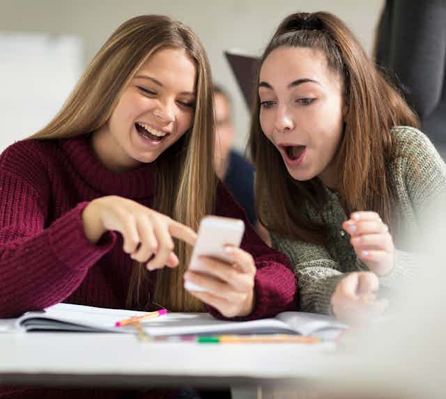 Two smiling female students look at a cell phone while seated at a desk in their class. They are in their teens or early 20s.