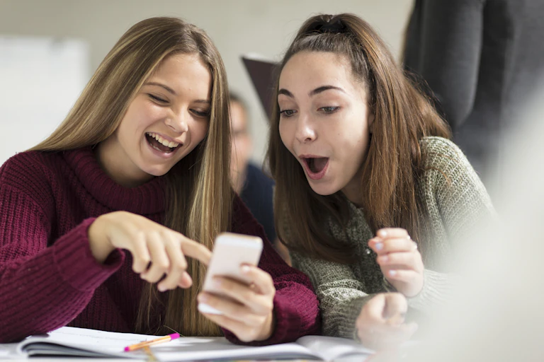 Two smiling female students look at a cell phone while seated at a desk in their class. They are in their teens or early 20s.
