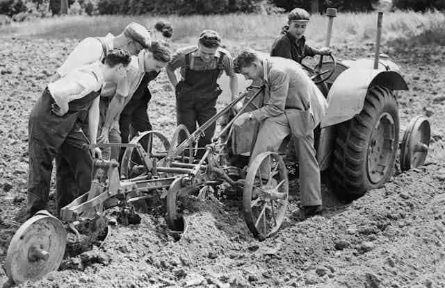 Group of men inspect a plough behind a tractor in a field