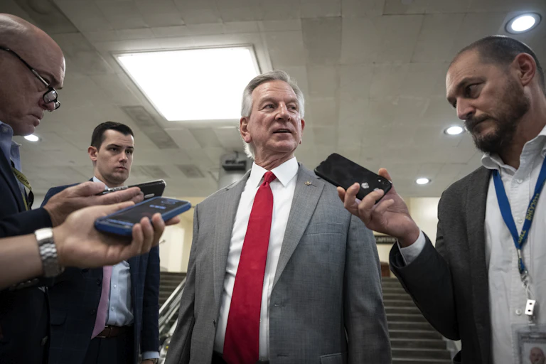A gray haired man wearing a gray suit jacket and red tie speaks into phones and reorders held by three male reporters.