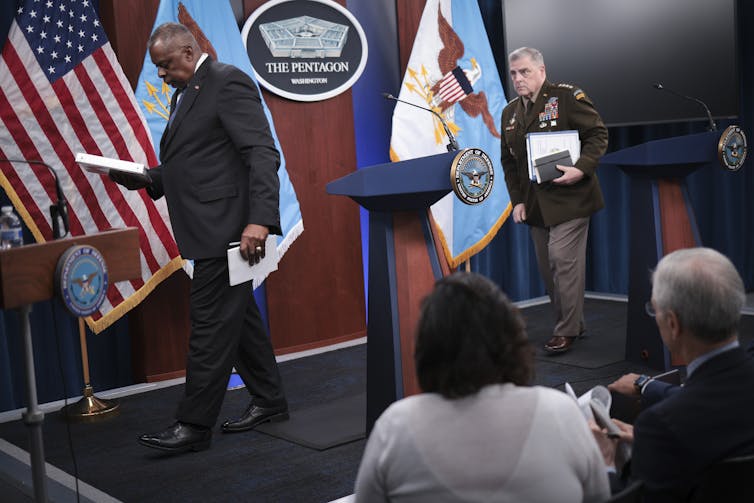 A man wearing a dark suit walks toward a lectern, carrying a white binder in his right hand and paper and a pen in his left. He is fallowed by a man wearing a brown military uniform, carrying papers clutched under his left arm.