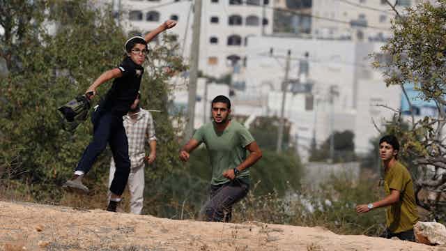 A boy with glasses on is seen about to hurl a stone toward the cameras. Other boys are around him.