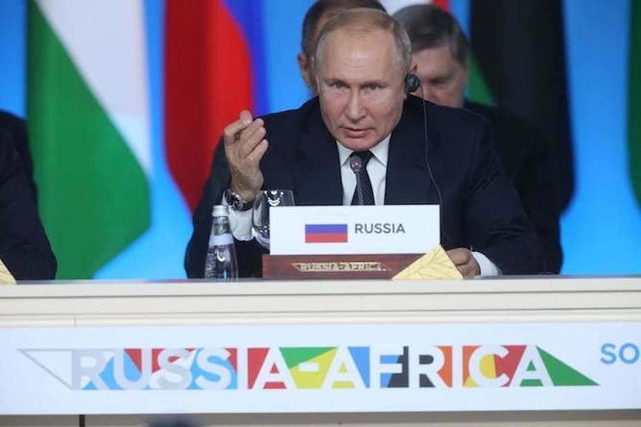 Un homme fait des gestes alors qu'il parle dans un microphone, assis. Devant lui, une étiquette portant l'inscription "Russie" est posée sur une table où sont inscrits les mots "Russie-Afrique".
