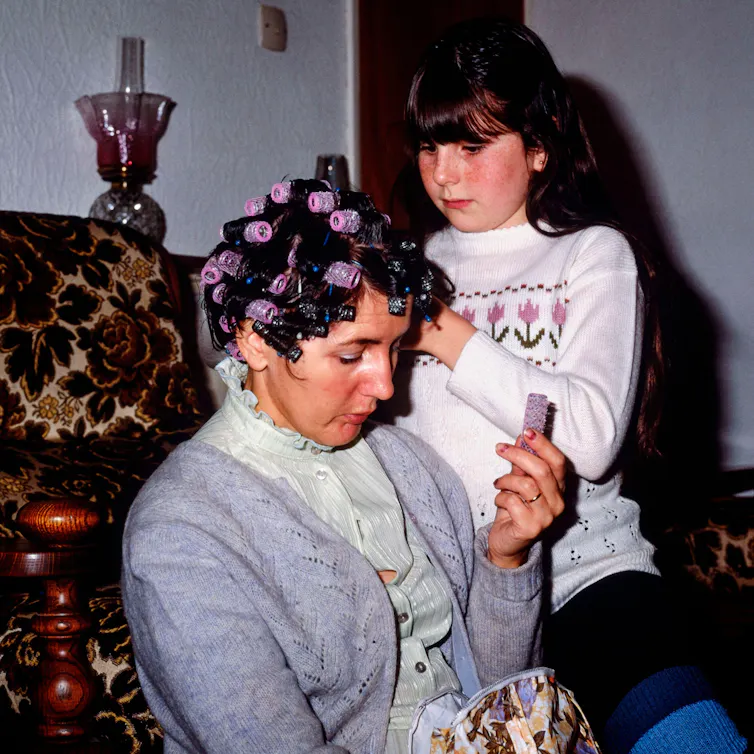 An archival colour photograph of a young girl putting curlers in her mother's hair.