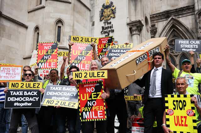 A group of anti-Ulez protesters with various signs calling to 'take back democracy'. One man is carrying a coffin for democracy