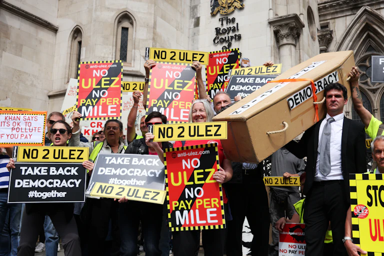 A group of anti-Ulez protesters with various signs calling to 'take back democracy'. One man is carrying a coffin for democracy