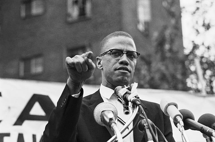 A man wearing glasses and a suit speaks in front of microphones at a rally.