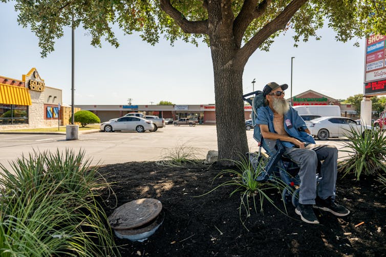 A man sits in a chair under a tree in a shopping center parking lot. He's wearing a baseball cap and his shirt is open to let the breeze in.