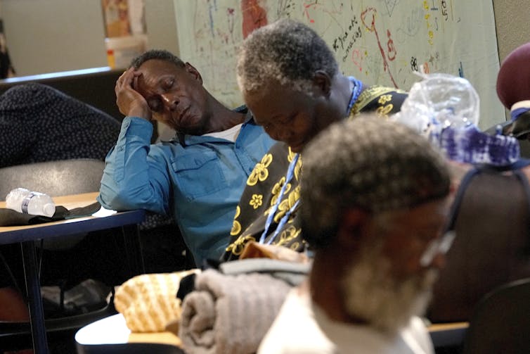 A man rests while propping his head up with his arm on a table. A woman dozes in a chair in a room with several other people.