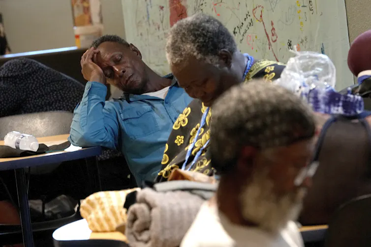 A man rests while propping his head up with his arm on a table. A woman dozes in a chair in a room with several other people.