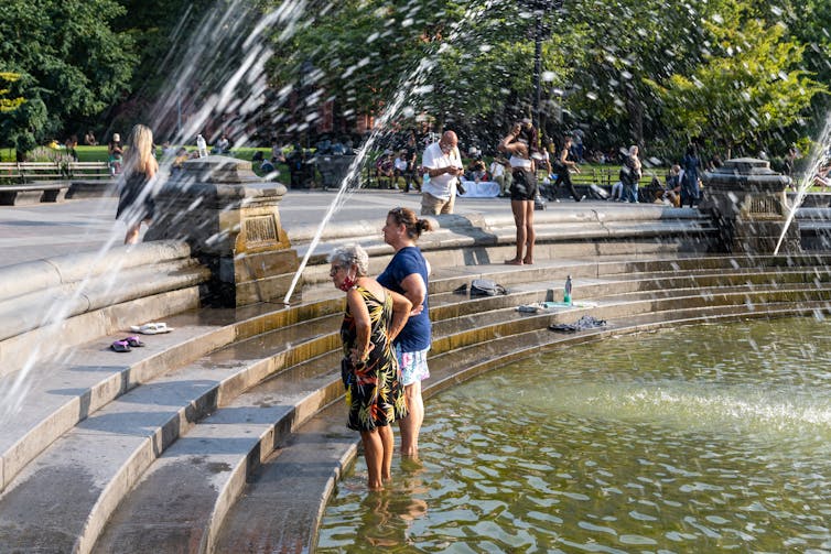 Two women stand in a large fountain in Washington Square park. It has steps designed for people to sit or walk down to the water.