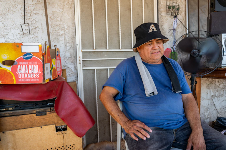 A man sits in front of a fan on his porch in Phoenix in July 2023, with a wet towel draped on his neck and a fan blowing from the side.