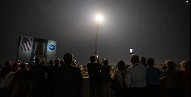 A crowd of people stand filming a rocket launch in front of a building with a NASA sign. 