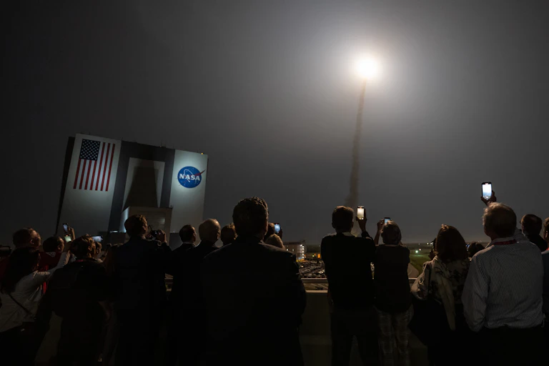 A crowd of people stand filming a rocket launch in front of a building with a NASA sign.