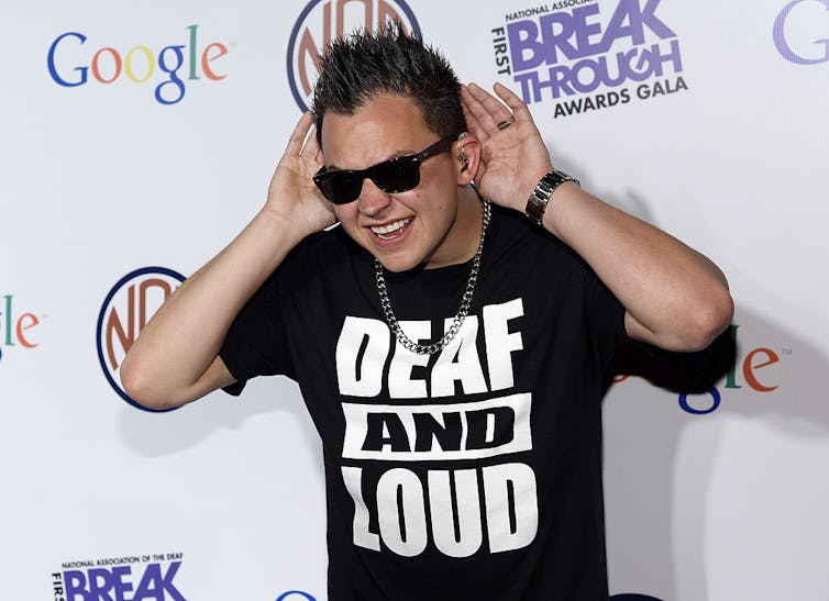 Man wearing sunglasses and a shirt that reads 'deaf and loud' holds his hands up to his ears.
