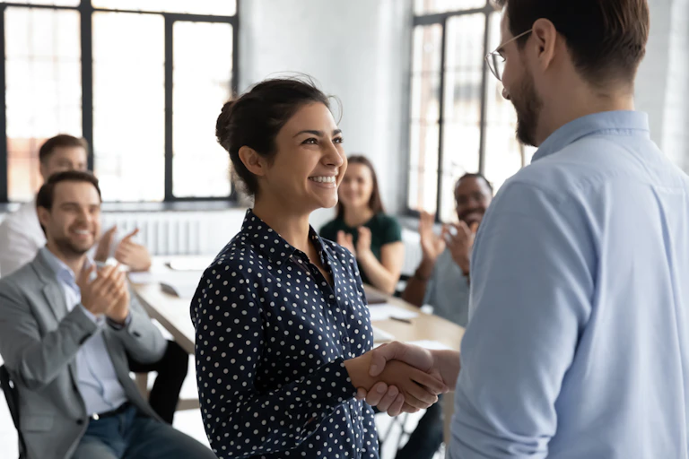 Smiling woman and man standing up shaking hands as table of seated men and women clap and smile.