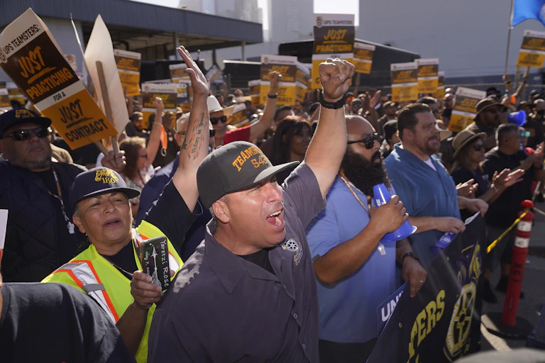 Workers, some holding UPS-branded signs that say 'just practicing' gather together to protest.
