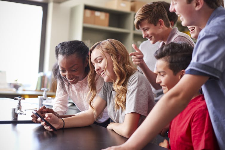 A group of kids in a classroom looks at a phone.