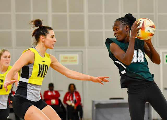 Two women face off on a netball court. An African woman holds the ball at shoulder height while another woman defends, arms outstretched.