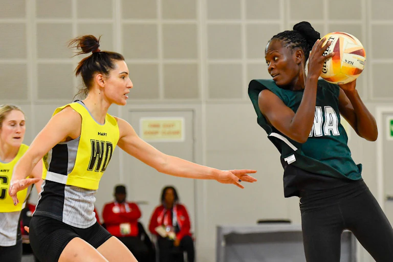 Two women face off on a netball court. An African woman holds the ball at shoulder height while another woman defends, arms outstretched.
