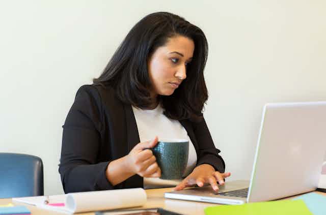 A dark-haired woman drinking from a mug and typing on a computer.