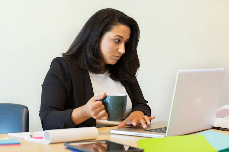 A dark-haired woman drinking from a mug and typing on a computer.